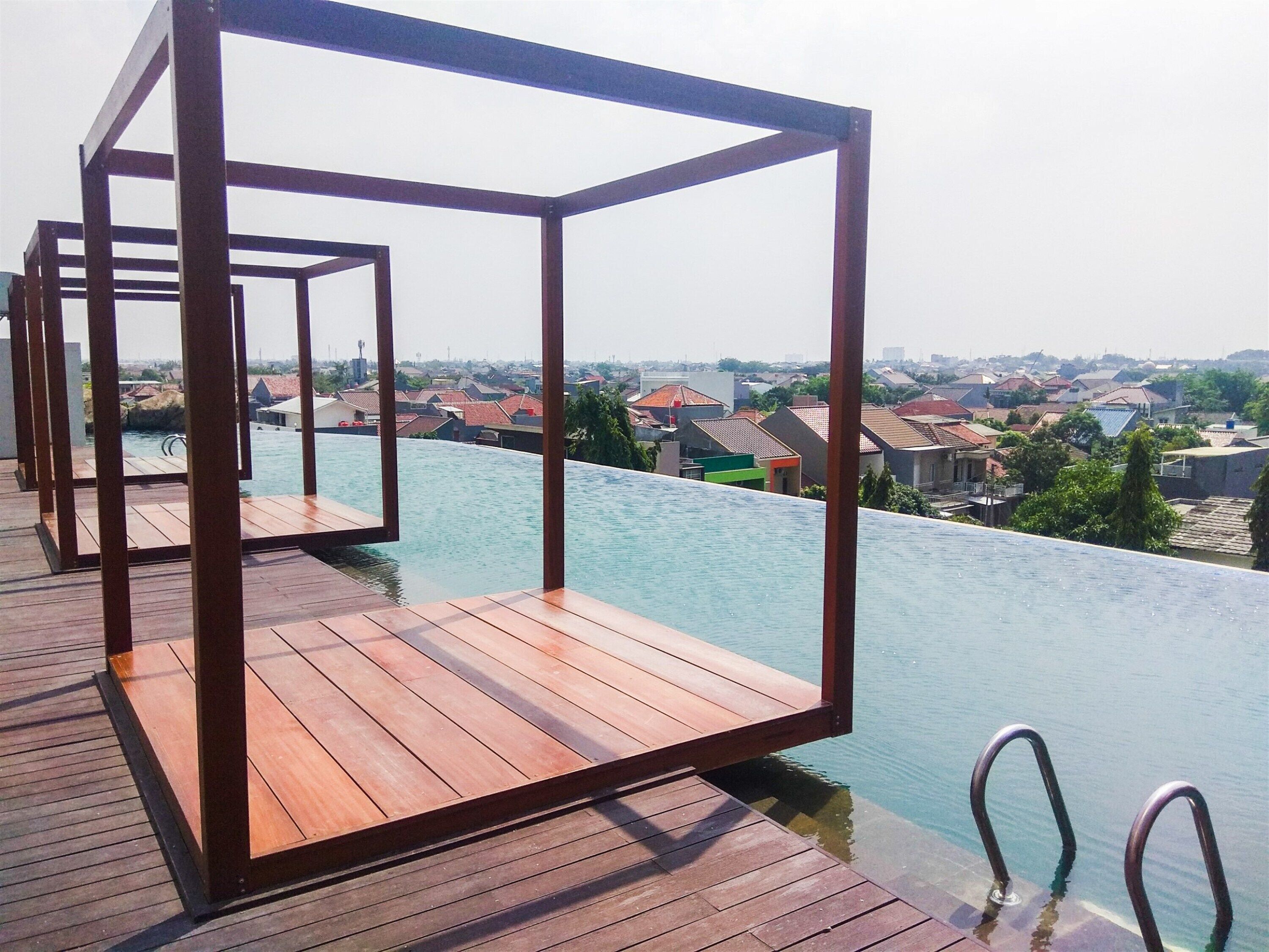 An infinity pool with a wooden deck and metal railings, overlooking a residential area.
