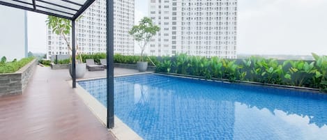 A rooftop swimming pool with a tiled floor, surrounded by greenery and a view of tall buildings.