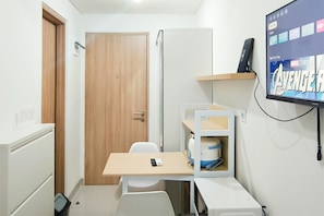 Una habitación pequeña con puerta de madera, un escritorio blanco con una silla, un estante con un televisor mostrando 'AVENGERS' en la pantalla y un electrodoméstico blanco.