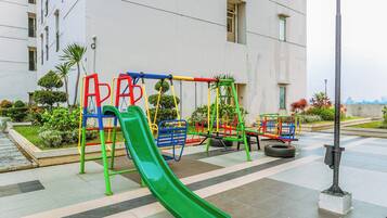 Um parquinho ao ar livre com escorrega verde, balanços e um balanço de pneu, cercado por um jardim bem cuidado e um edifício ao fundo.