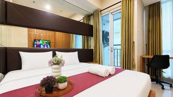 A hotel room with a large bed, a TV, a balcony with curtains, and a desk with a chair.