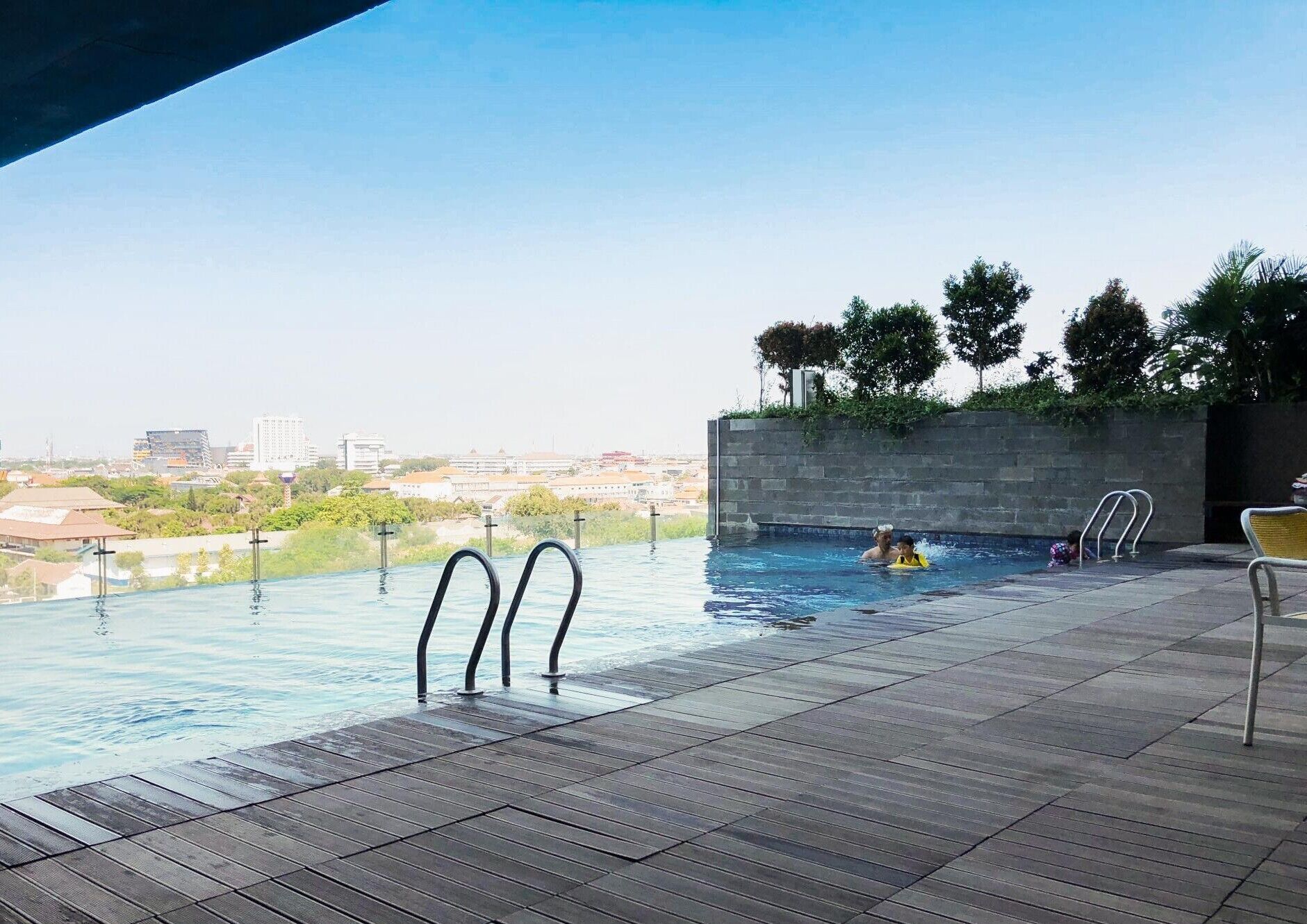 An outdoor infinity pool with a city view, a wooden deck, and a stone wall with greenery.