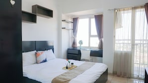 A hotel room with a bed, a desk, a chair, a window with curtains, and a view of the outside. - Cozy And Relaxing Studio At Apartment Margonda Residence 5 (Depok)