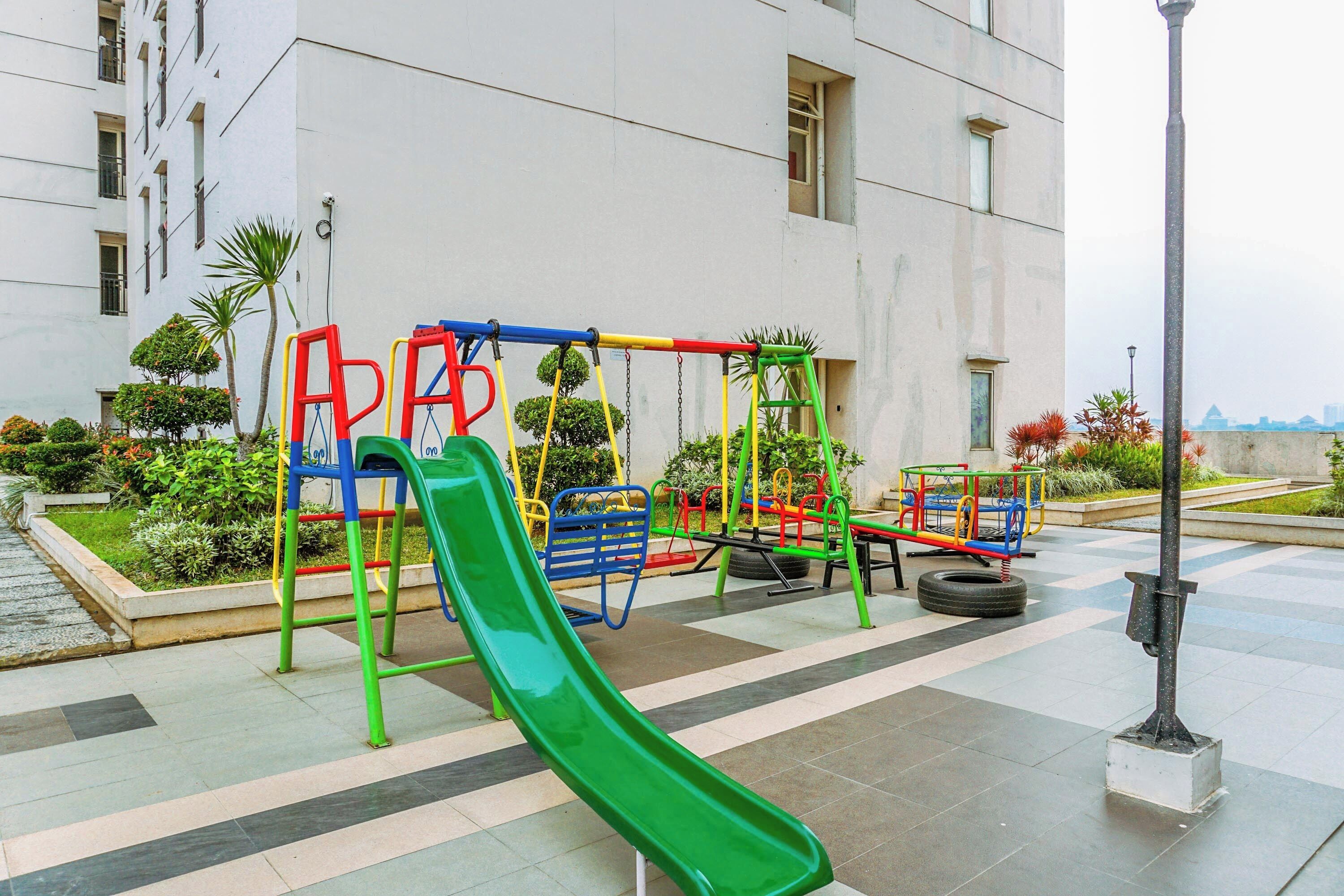 An outdoor playground with a green slide, swings, and a tire swing, surrounded by a landscaped garden and a building in the background.