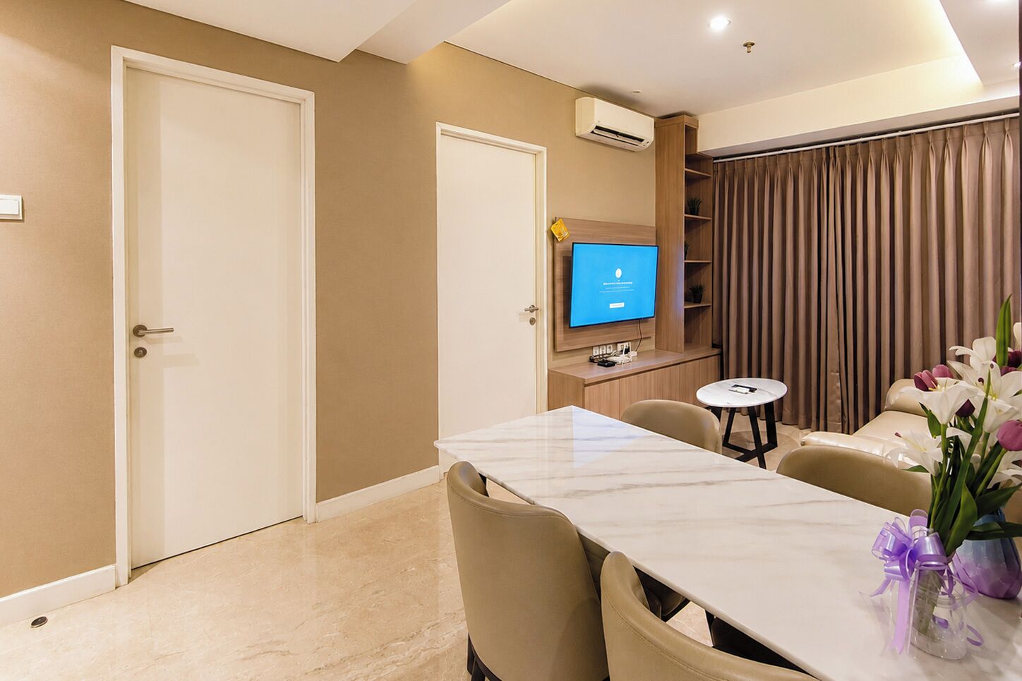 A hotel room with a dining table, chairs, a television, and a small round table with a vase of flowers.
