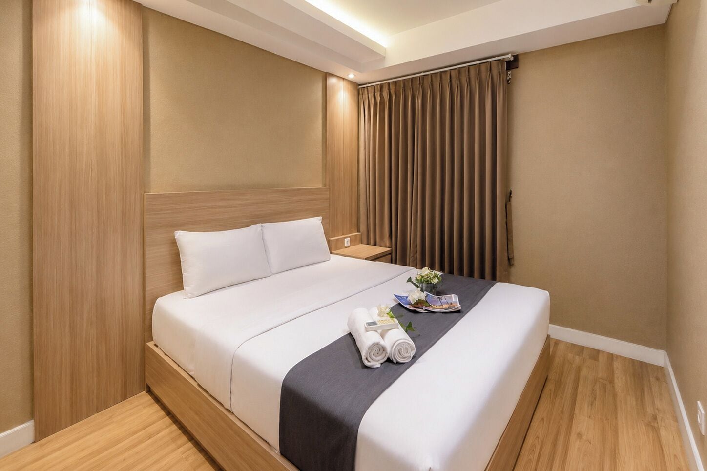 A hotel room with a bed, wooden headboard, curtains, and a small table with a flower arrangement.