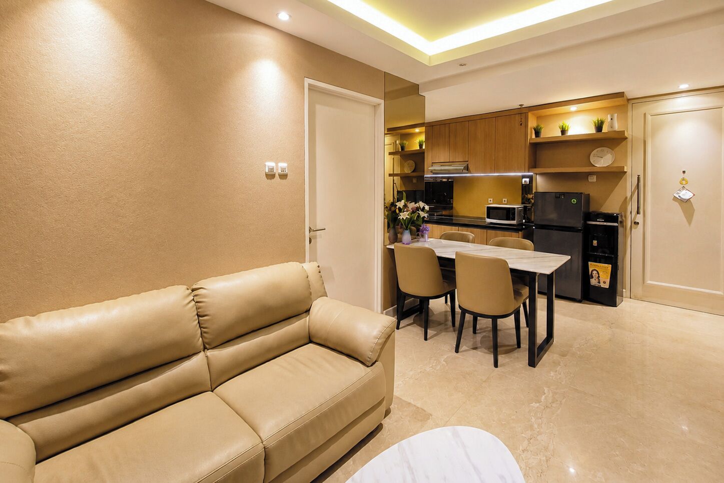 A modern living room with a beige leather sofa, a dining table with chairs, and a kitchen area with wooden cabinets and appliances.
