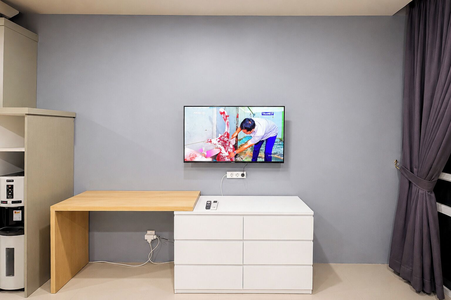Habitación con televisión de pantalla plana en la pared, un escritorio de madera, un mueble de almacenamiento blanco y una cortina gris.