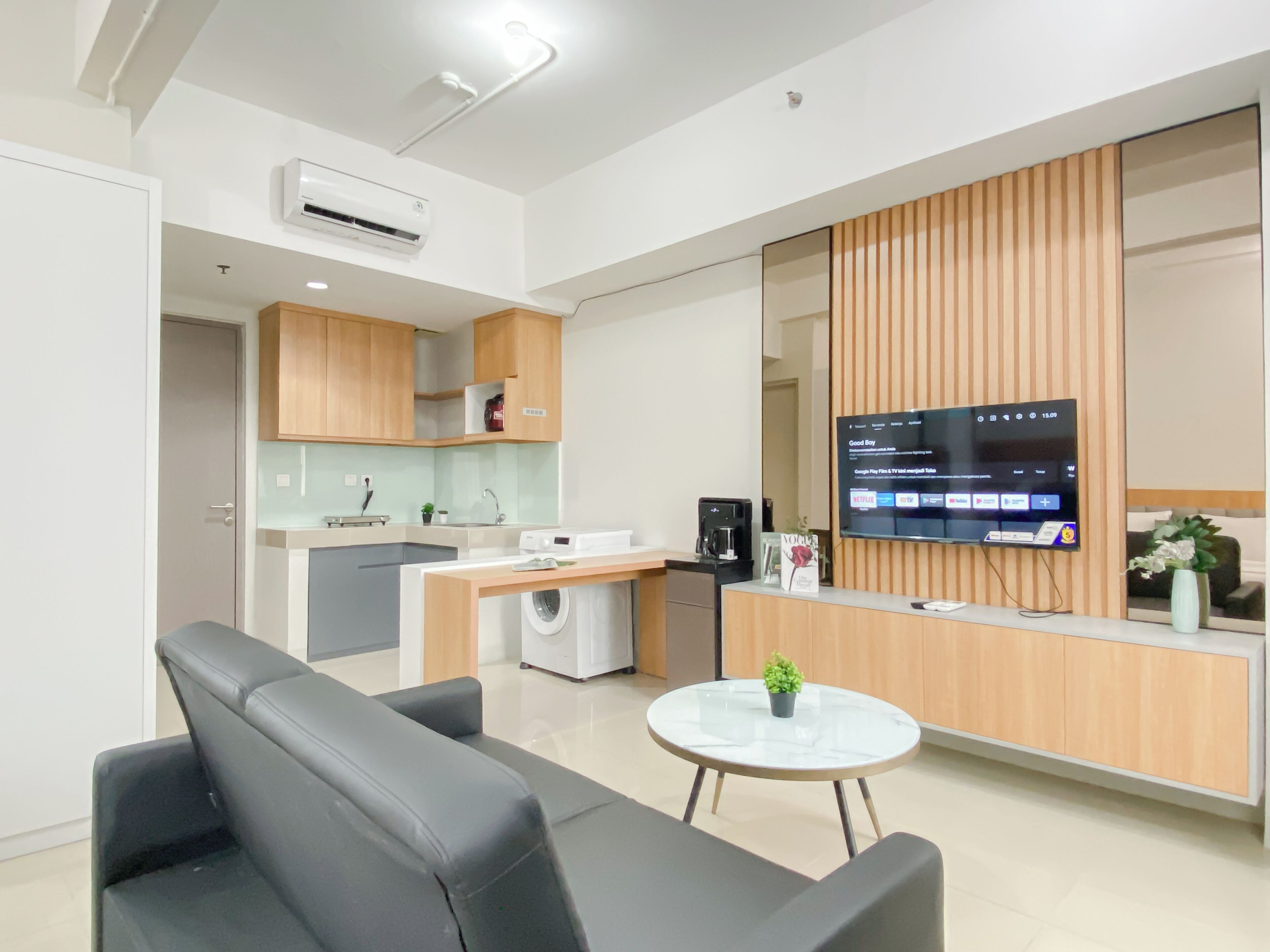 A modern living room with a sofa, a coffee table, a TV, and a kitchen area with wooden cabinets and appliances.