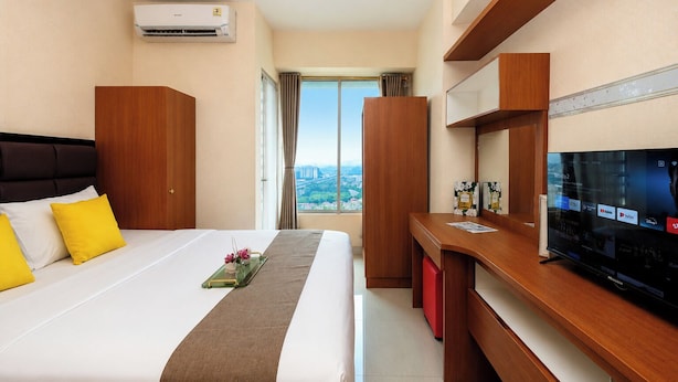 A hotel room with a bed, a desk, a television, and a view of a cityscape. - Comfortable 1Br Without Living Room At Grand Kamala Lagoon Apartment (Bekasi)