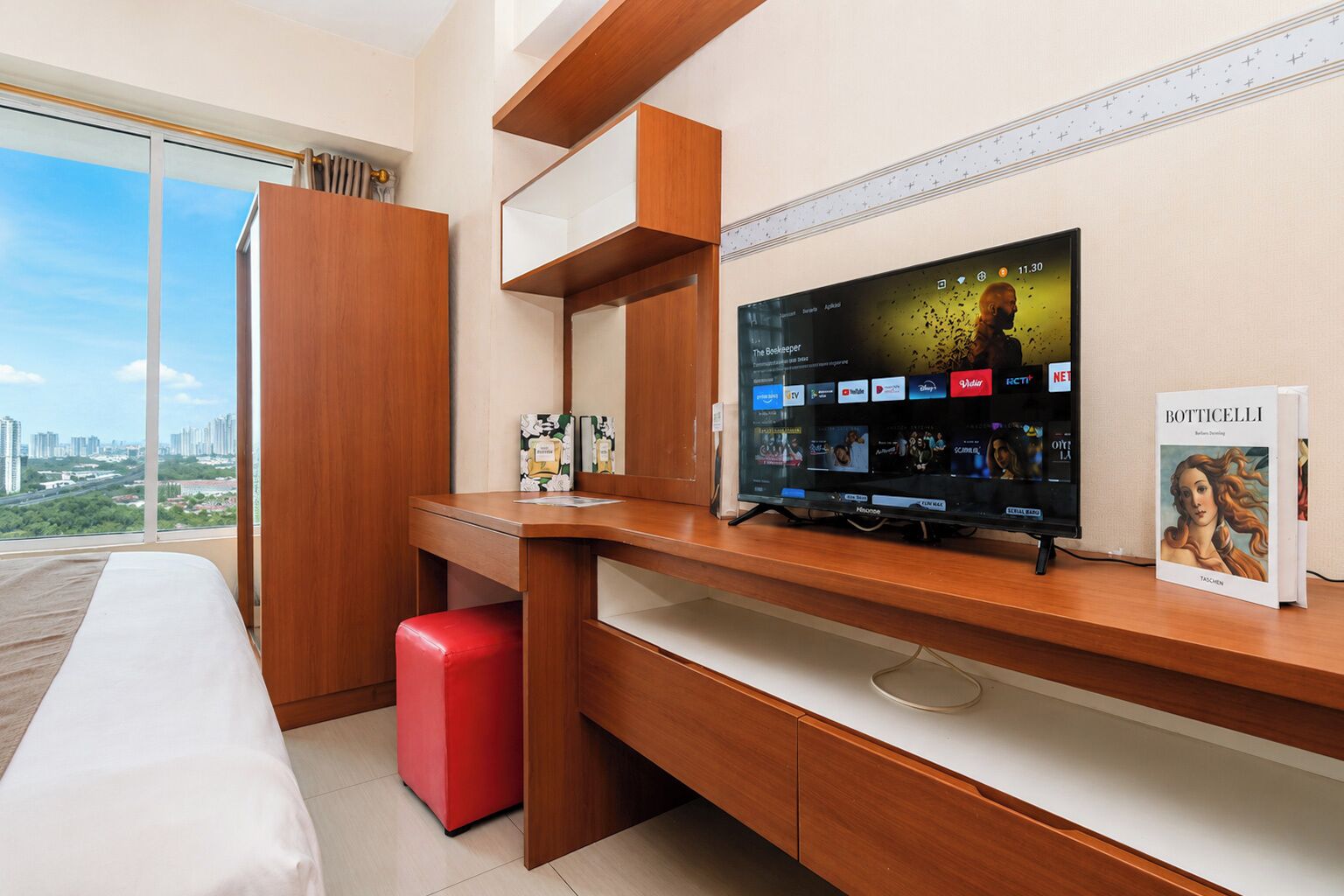 Habitación de hotel con un televisor de pantalla plana grande, un escritorio de madera, un reposapiés rojo y un ventanal con vistas a la ciudad.