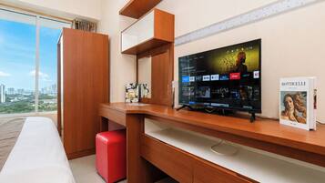 Habitación de hotel con un televisor de pantalla plana grande, un escritorio de madera, un reposapiés rojo y un ventanal con vistas a la ciudad.