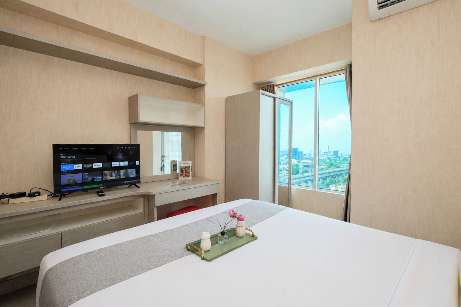 Quarto de hotel com cama, televisão, escrivaninha e janela com vista para a cidade.