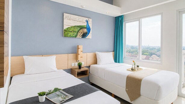 Una camera d'albergo con due letti, un quadro di un pavone, vista sulla città e una finestra con tende.