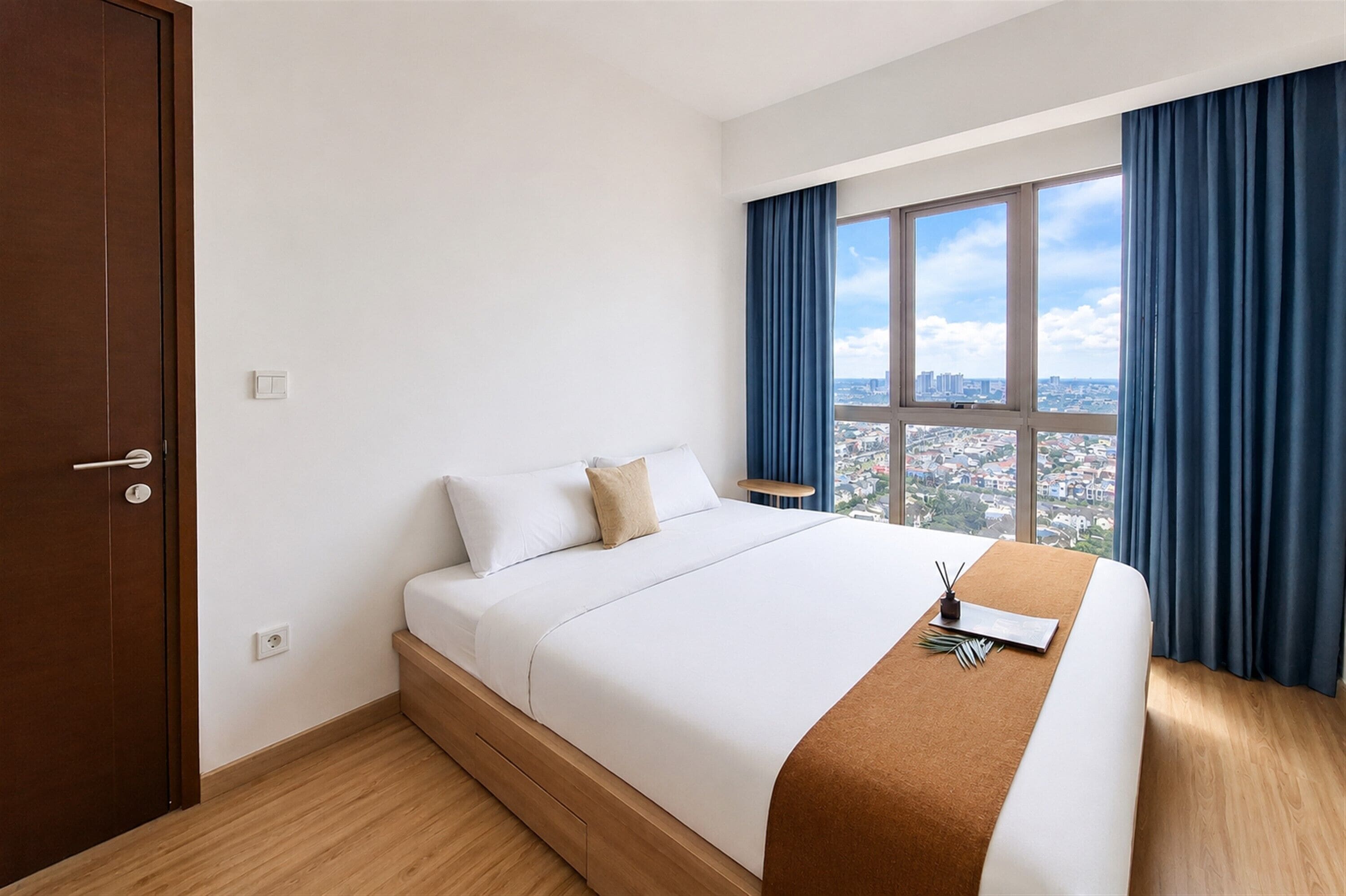 A hotel room with a bed, a window with curtains, a view of a cityscape, and a wooden floor.