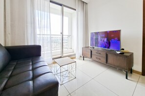 A living room with a leather sofa, a television on a wooden stand, and a balcony with a view of the city. - Homey And Well Located 1Br At Apartment M-Town Signature (Tangerang)
