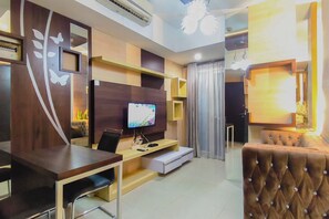 A modern living room with a flat-screen TV, a brown tufted sofa, a wooden desk, and a built-in entertainment unit. - Well Furnished 2Br At Apartment Tamansari Papilio (Surabaya)