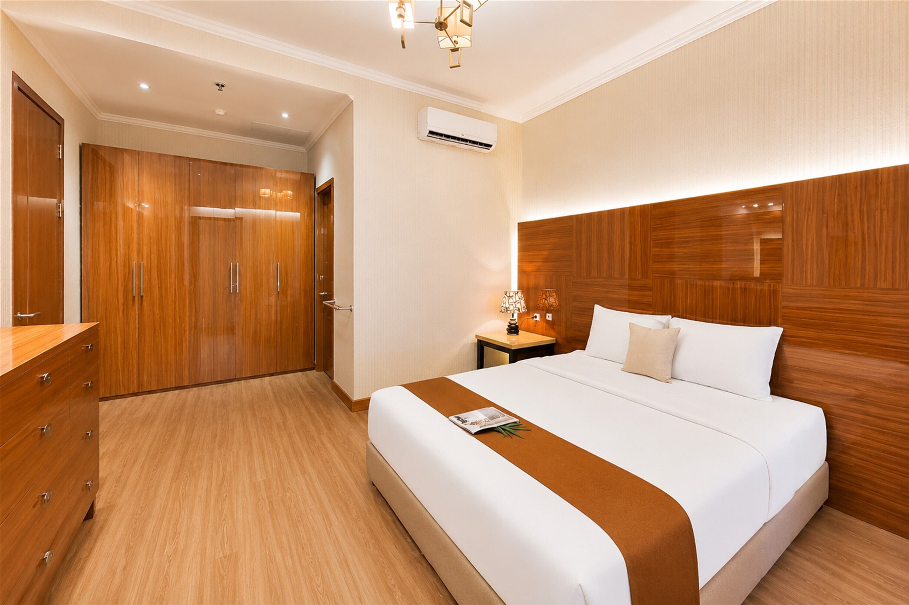 A hotel room with a wooden wardrobe, a bed with a white and brown bedspread, a nightstand with a lamp, and an air conditioning unit on the wall.