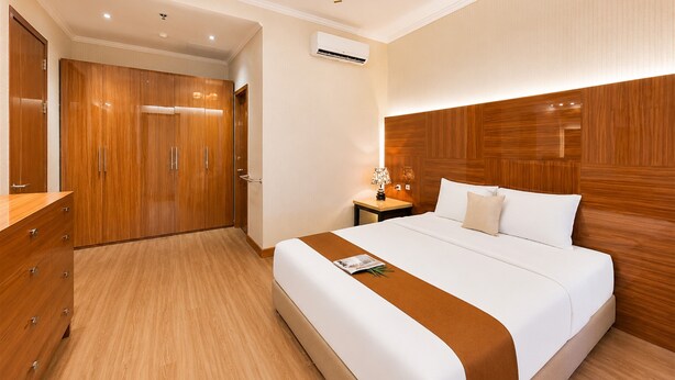 A hotel room with a wooden wardrobe, a bed with a white and brown bedspread, a nightstand with a lamp, and an air conditioning unit on the wall. - Comfortable 3Br Apartment At Great Western (Tangerang)