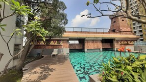 A swimming pool with a wooden deck, surrounded by greenery and a building with a decorative facade. - Comfortable 3Br Apartment At Great Western (Tangerang)