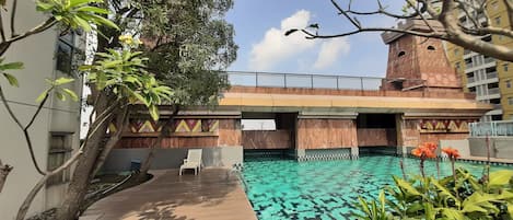 A swimming pool with a wooden deck, surrounded by greenery and a building with a decorative facade.