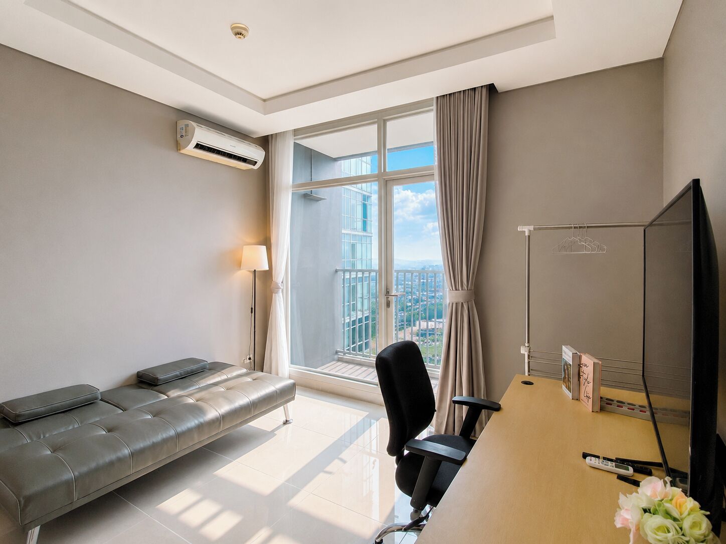 A modern hotel room with a large window, a leather sofa, a desk with a chair, and an air conditioning unit.