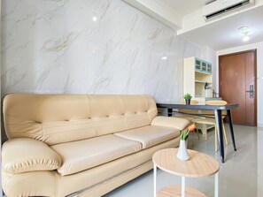 A modern living room with a beige sofa, a small round wooden table with a vase, and a dining area with a table and chairs. - Comfortable 2Br At Apartment Signature Park Grande (Jakarta)
