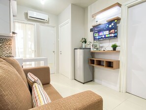 A modern living room with a sofa, a flat-screen TV mounted on a wall, a refrigerator, and a small shelf with decorative items. - Nice Homey 2Br At Apartment Green Pramuka City (Jakarta)