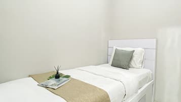 Una cama individual con cabecera blanca, almohada gris, manta color beige y una mesita con una revista y un difusor de cañas.