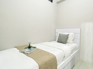A single bed with a white headboard, a grey pillow, a beige throw blanket, and a small table with a magazine and a reed diffuser. - Nice Homey 2Br At Apartment Green Pramuka City (Jakarta)