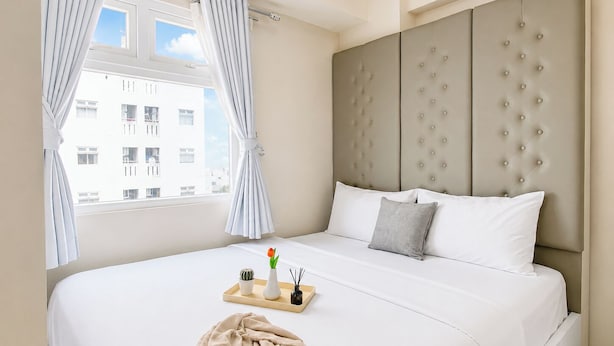 Una camera d'albergo con un letto grande, una finestra con tende e vista sugli edifici, e un comodino con un vassoio.