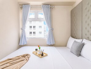 A hotel room with a bed, a window with curtains, a view of buildings, and a bedside table with a tray. - Nice Homey 2Br At Apartment Green Pramuka City (Jakarta)