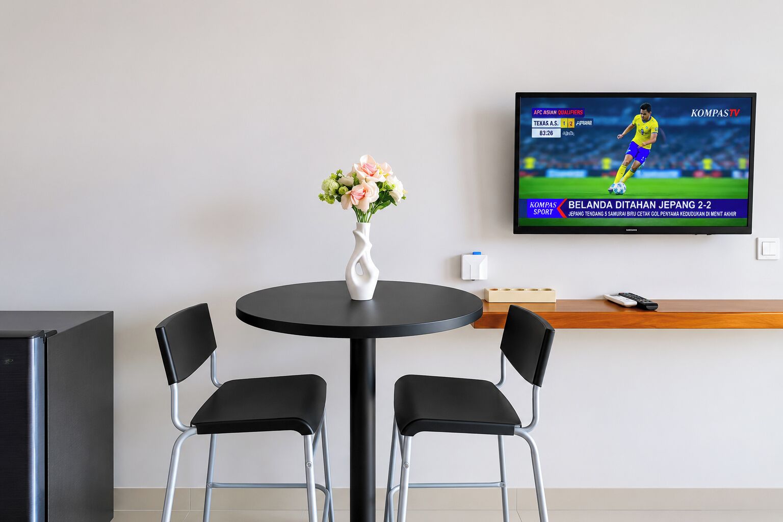 A hotel room with a round table, two chairs, a vase with flowers, a television displaying a sports event, and a minibar.