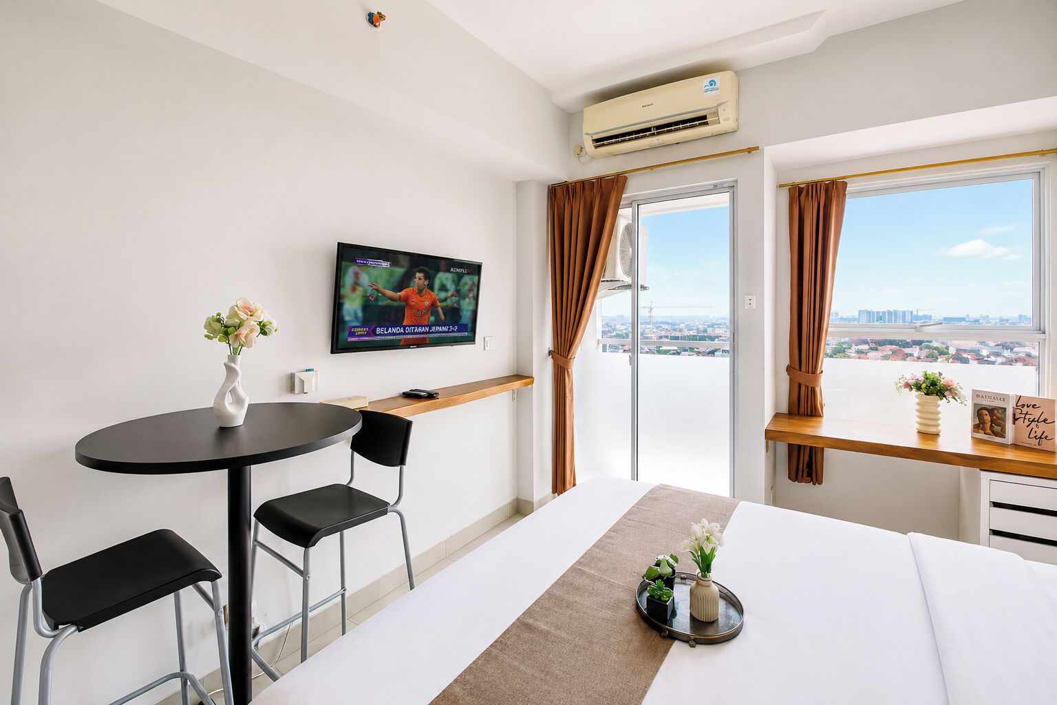 A hotel room with a bed, a TV, a table with chairs, a window with curtains, and a view of the city.