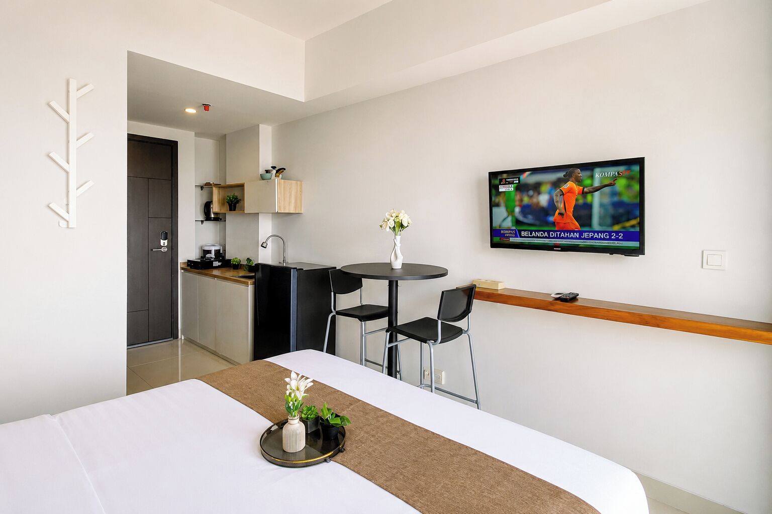 Een hotelkamer met een bed, een eettafeltje, een aan de muur bevestigde televisie en een kitchenette op de achtergrond.