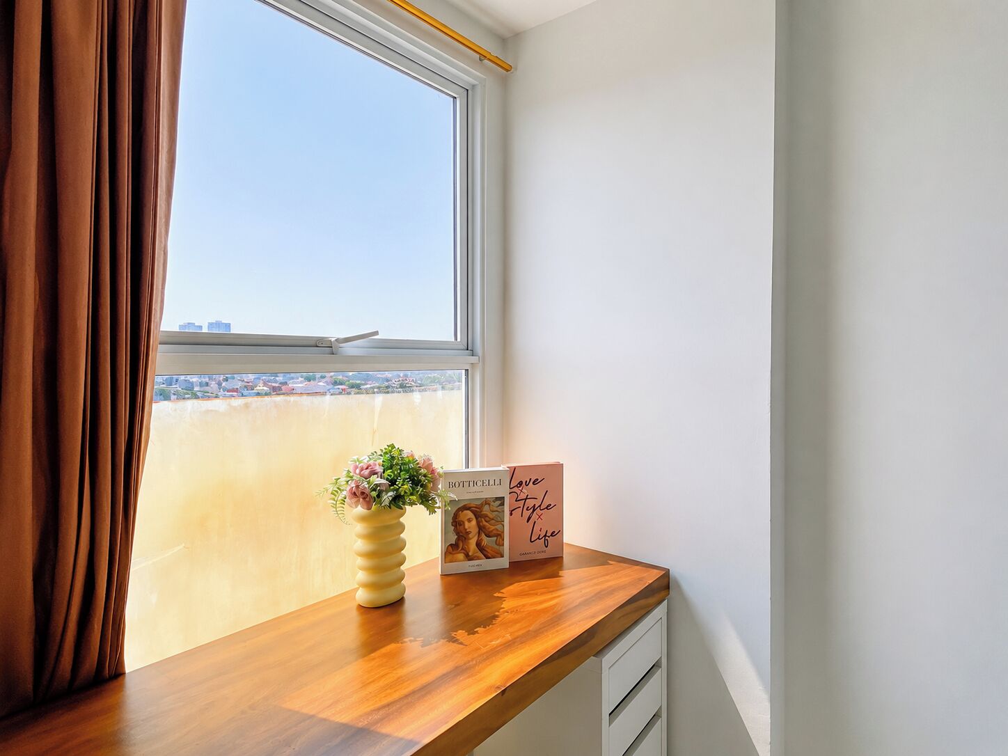A window with a view of a cityscape, a wooden desk with a vase of flowers, and a framed picture with text.