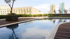 A swimming pool with a wooden deck, surrounded by greenery and modern buildings in the background. - Bright Minimalist 2Br At Apartment Elpis Residence (Jakarta)