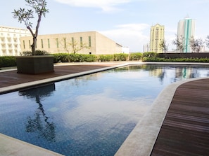 A swimming pool with a wooden deck, surrounded by greenery and modern buildings in the background. - Bright Minimalist 2Br At Apartment Elpis Residence (Jakarta)