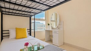 A hotel room with a bed, a bedside table with a mirror, a window with a view of buildings, and a small desk with a chair. - Bright Minimalist 2Br At Apartment Elpis Residence (Jakarta)