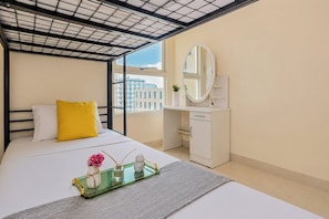 A hotel room with a bed, a bedside table with a mirror, a window with a view of buildings, and a small desk with a chair. - Bright Minimalist 2Br At Apartment Elpis Residence (Jakarta)
