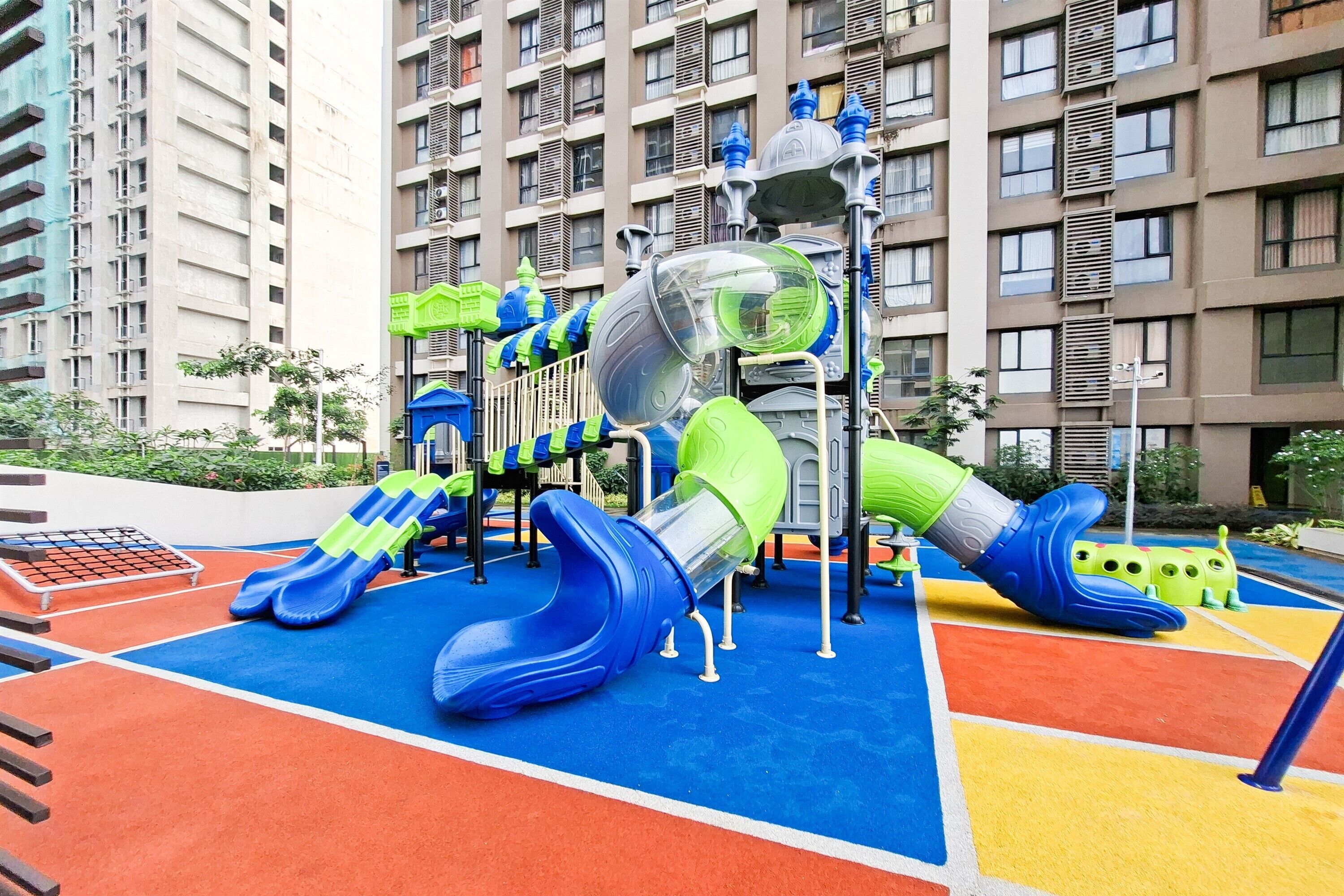 A playground with slides and climbing structures, surrounded by a colorful play area and tall residential buildings in the background.