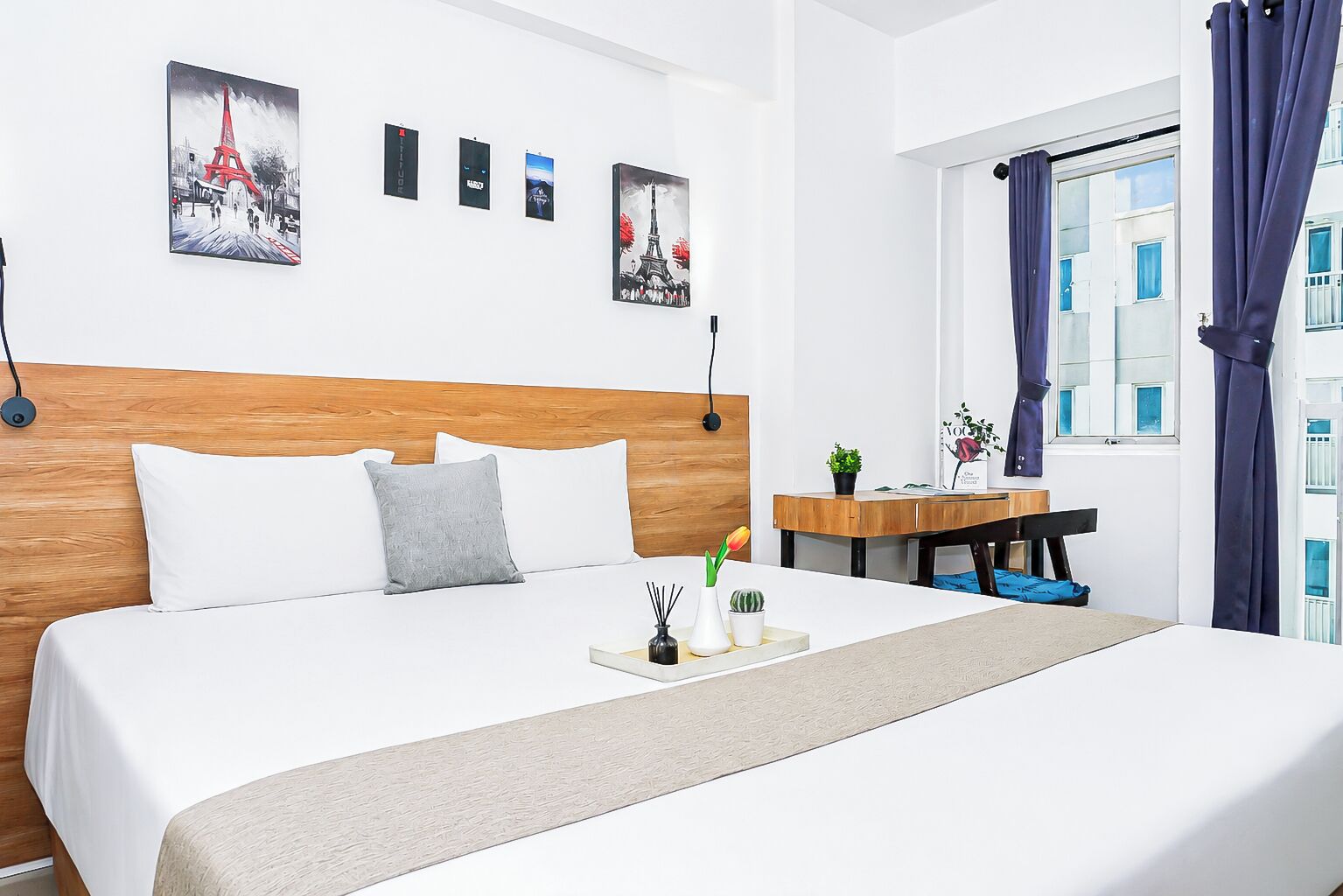 A hotel room with a wooden headboard, a bed with white linens, a bedside table with a plant, and artwork on the wall.
