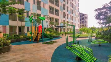 Un parque infantil con toboganes y equipamiento de ejercicio, rodeado por un edificio con balcones y un jardín.