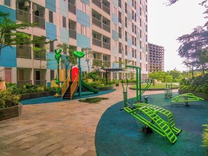 A playground with slides and exercise equipment, surrounded by a building with balconies and a garden. - Best Deal Studio Apartment At Grand Sentraland Karawang (Karawang)