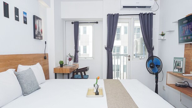 Hotellihuone, jossa on sänky, työpöytä, tuoli, ikkuna verhoilla, lattiatuuletin ja seinälle asennettu televisio.