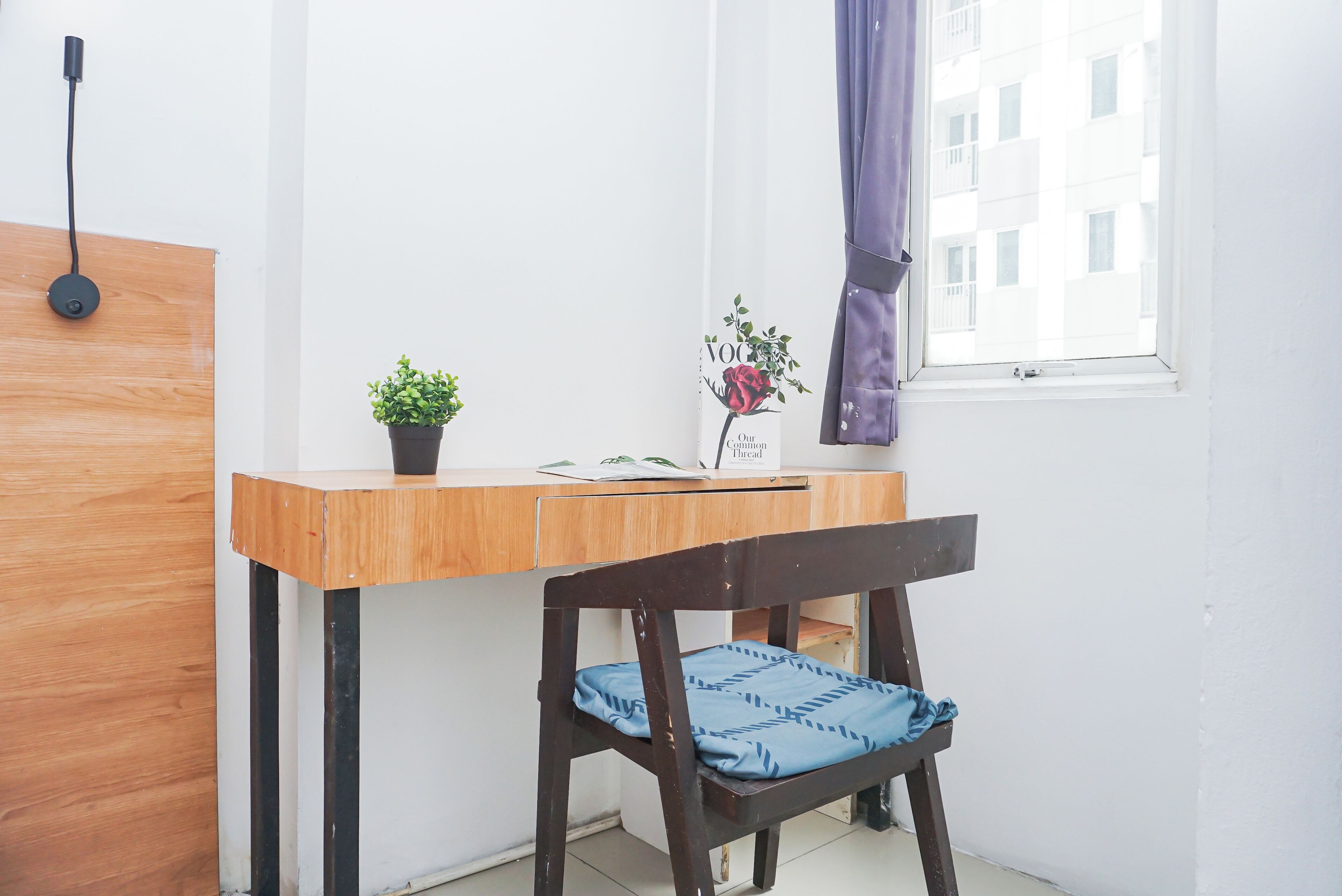 A small, tidy room with a wooden desk, a chair, a potted plant, and a window with a curtain.