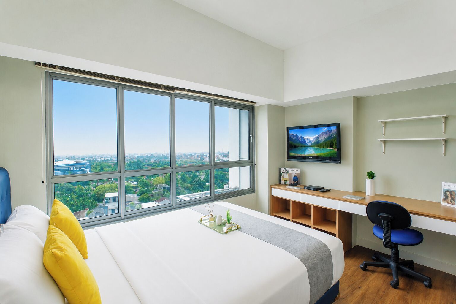 A hotel room with a large window, a bed, a desk with a computer, and a TV.