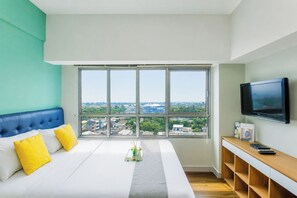 A hotel room with a large window, a bed with white and yellow bedding, a blue headboard, a wooden console table, and a flat-screen TV. - Neat And Nice Studio Room At Apartment Springlake Summarecon Bekasi (Bekasi)