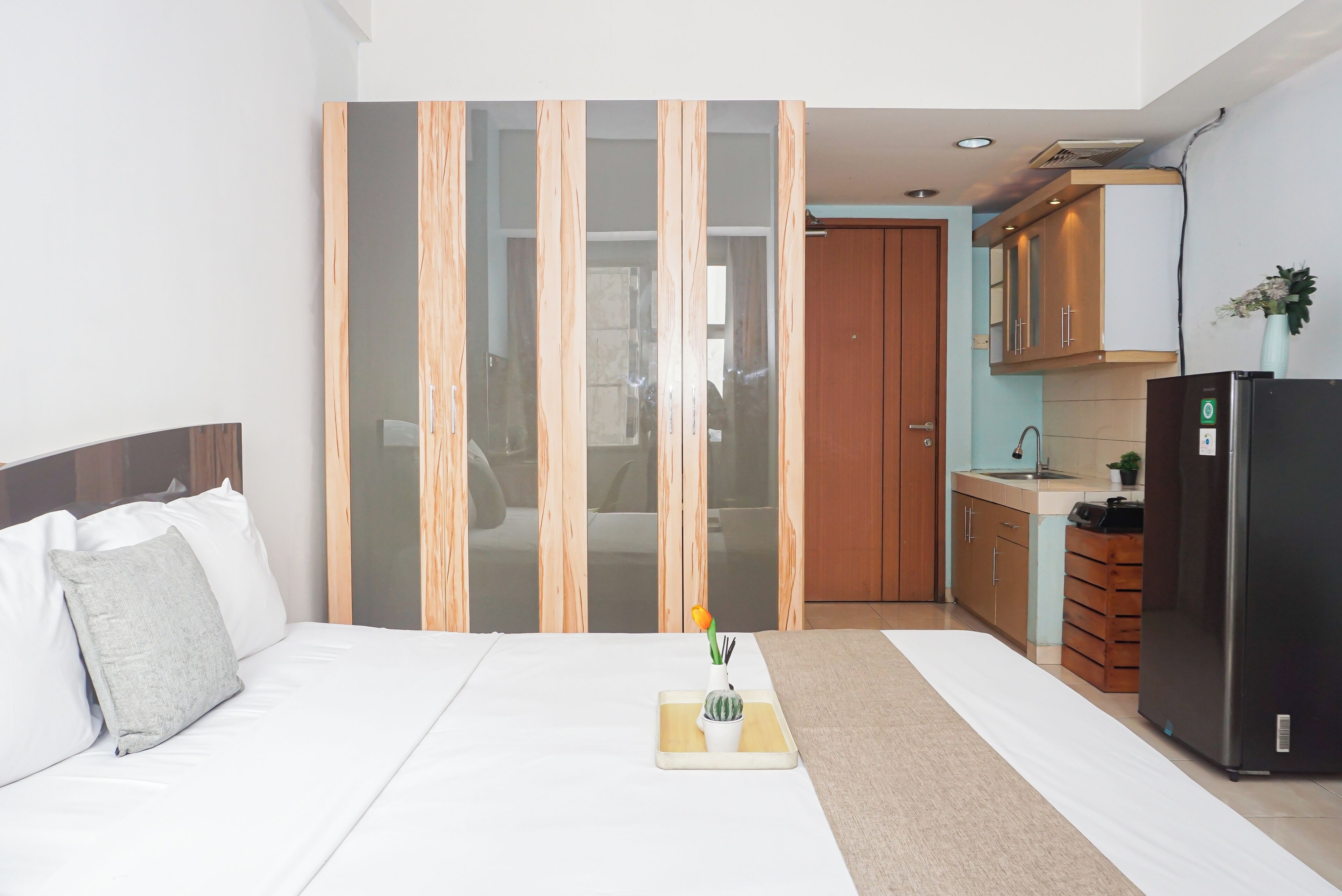 A bedroom with a bed, a bedside table with a vase and a plant, and a view into a kitchen area with cabinets and a refrigerator.