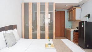 A bedroom with a bed, a bedside table with a vase and a plant, and a view into a kitchen area with cabinets and a refrigerator. - Compact Minimalist Studio At Apartment Margonda Residence 3 (Depok)